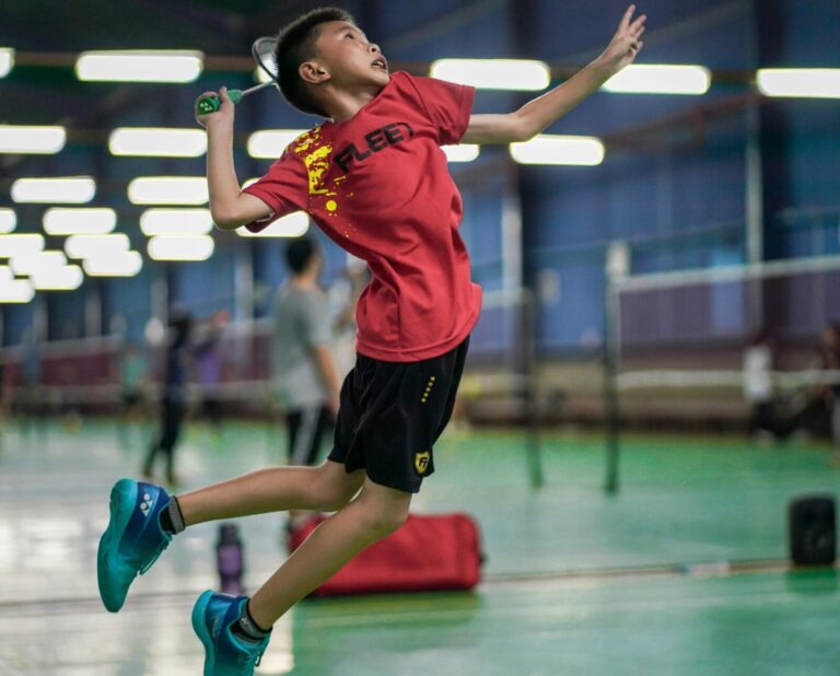 A young boy enthusiastically jumps to hit a badminton shuttlecock inside a sports hall.